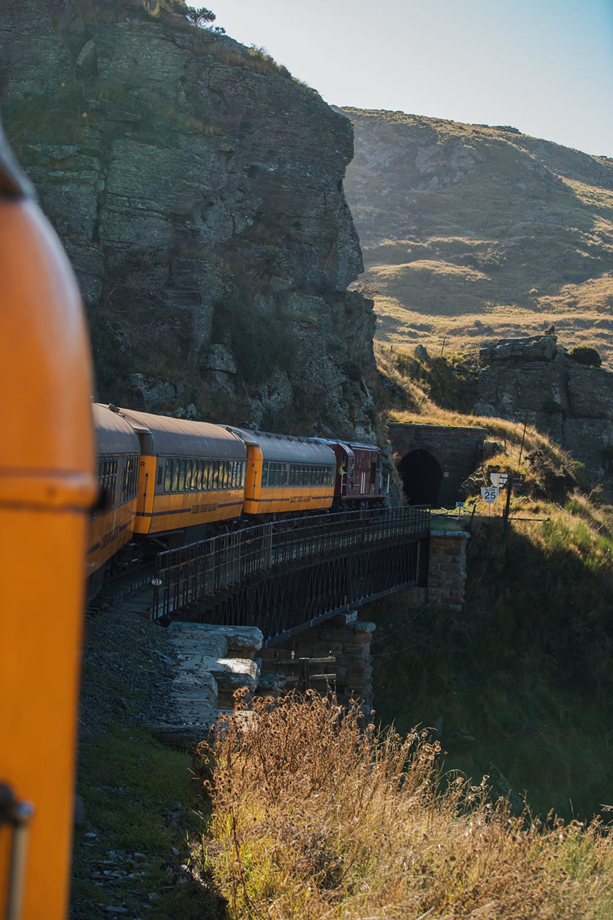 A yellow Taieri Gorge train passenger carriage crosses a bridge toward a mountain tunnel.