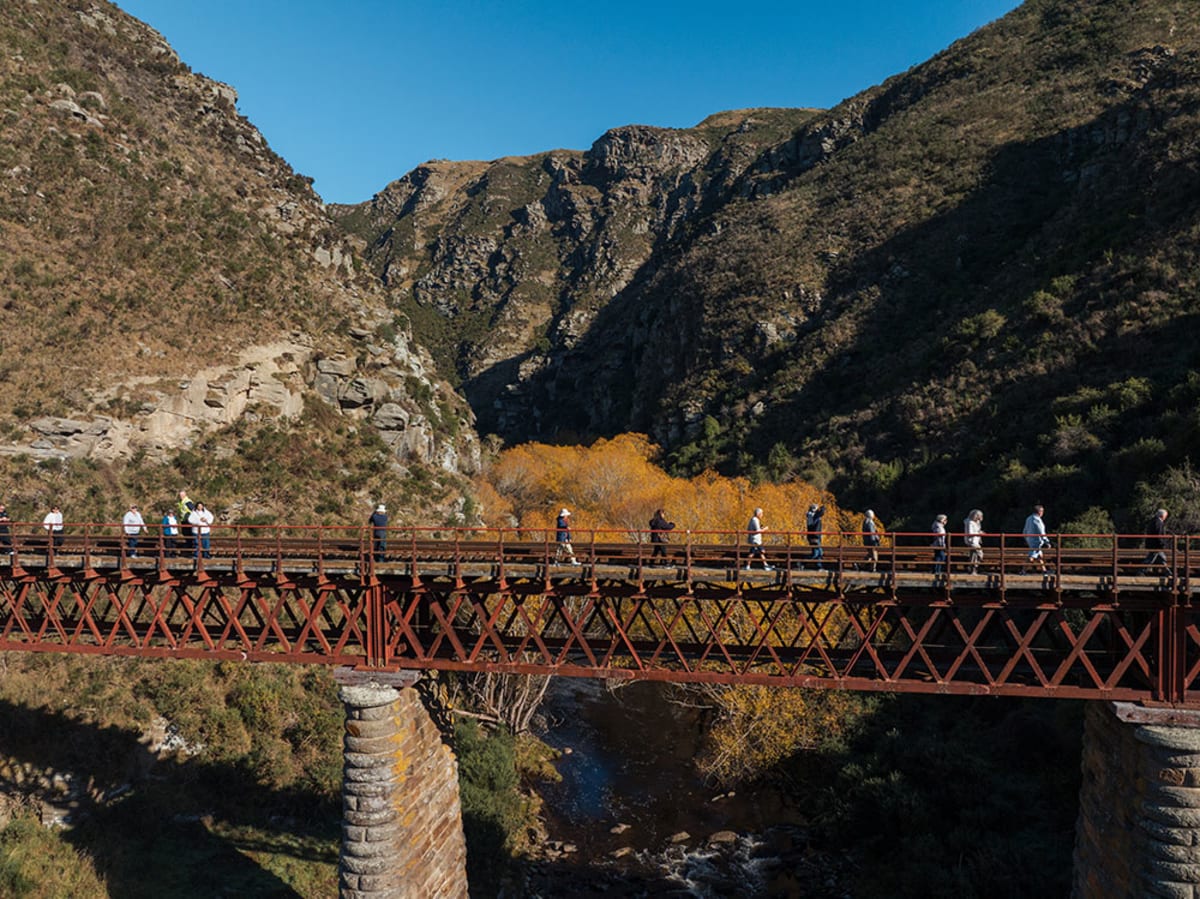 A group of people walks across the Wingaitui bridge over the Taieri river, surrounded by rugged hills and autumn trees.