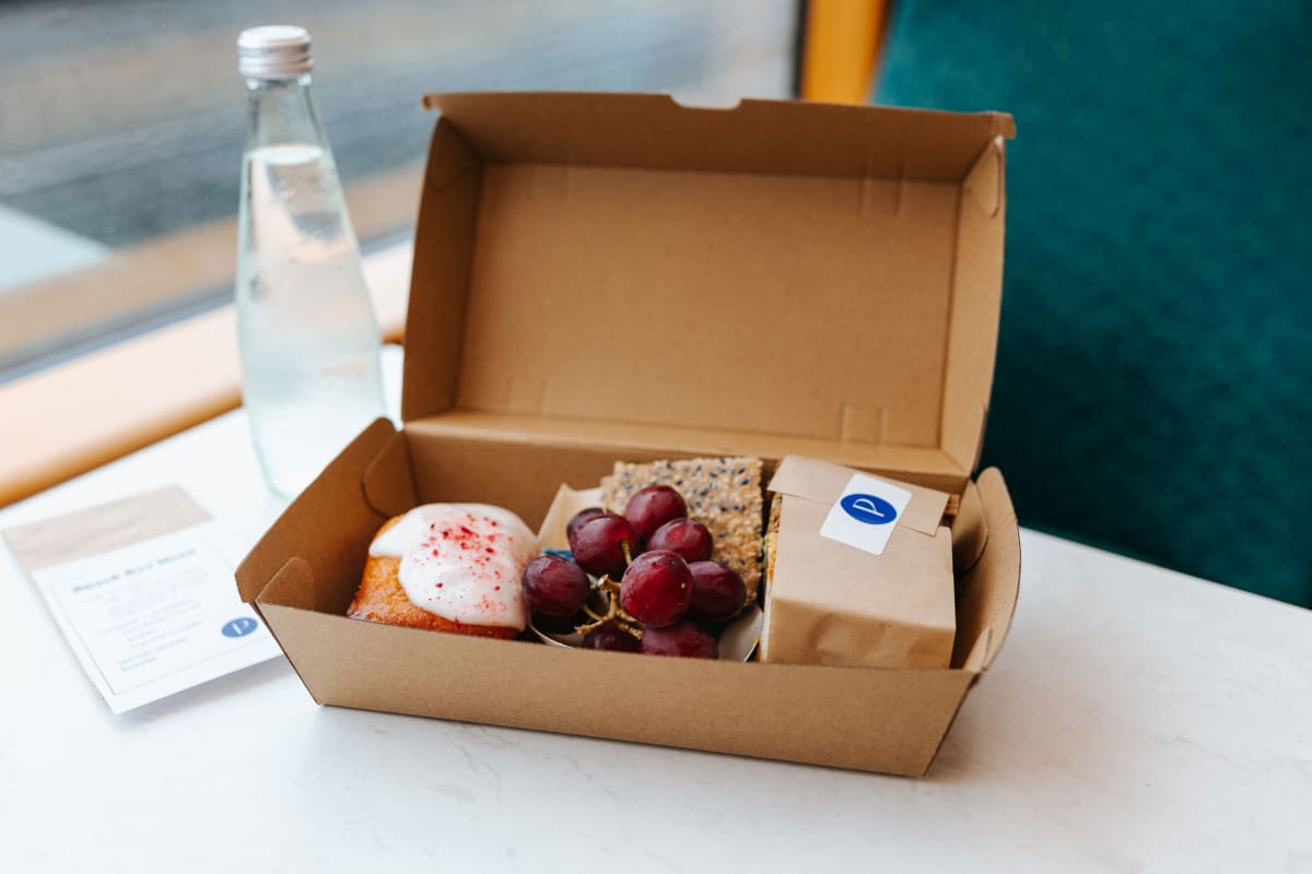 An open cardboard lunch box containing a frosted pastry, a bunch of grapes, crackers, and a wrapped item next to a bottled water aboard the Taieri Gorge Train.