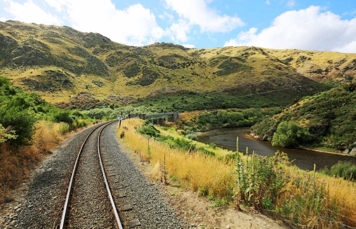 A curving heritage Dunedin railway track leads through the Taieri Gorge valley alongside the Taieri river, backed by rolling hills and a bridge.