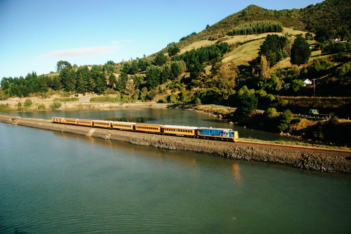 The Seasider train travels along a narrow causeway beside a body of water within Dunedin with a rolling green hill in the background.