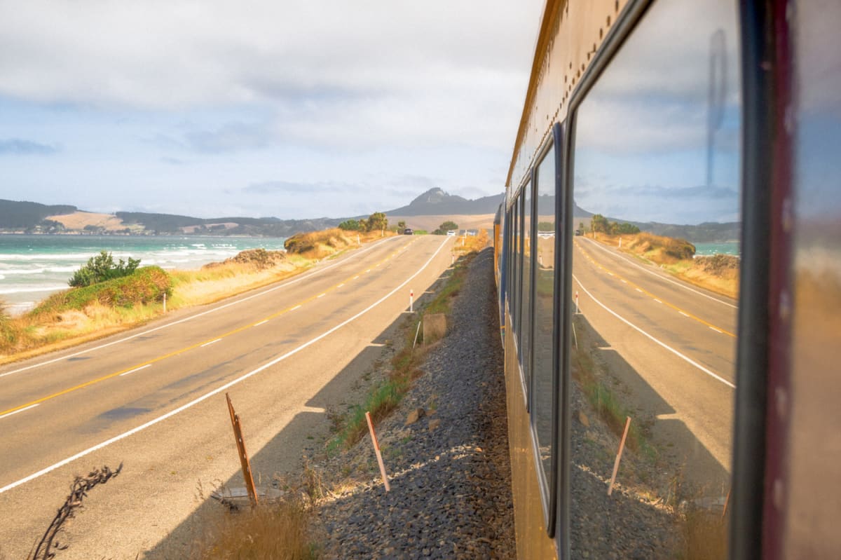 A NZ coastal road and distant hill are reflected in the windows of The Seasider train.