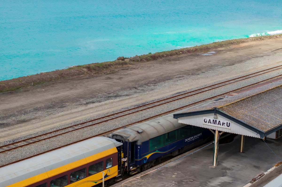 A Dunedin Railway Station passenger train waits at the Oamaru, NZ station platform beside coastal railway tracks.