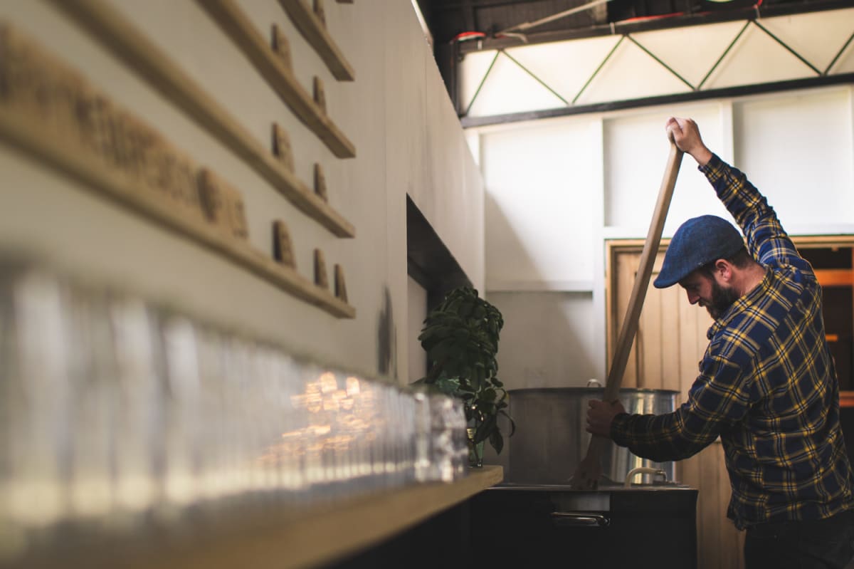 A man wearing a plaid shirt and flat cap uses a large wooden paddle to stir a large metal vat at Arc brewery.