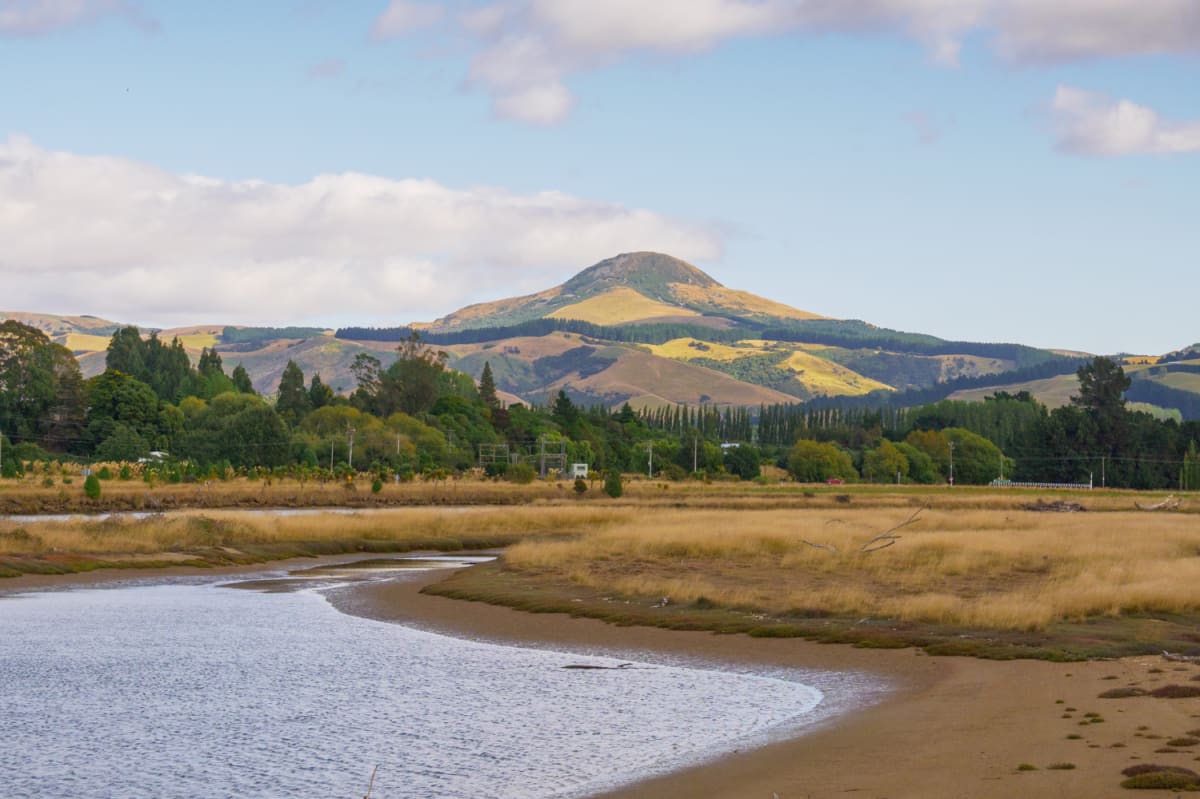 A winding tidal river flows through golden marshlands toward a large green hill under a partly cloudy sky.