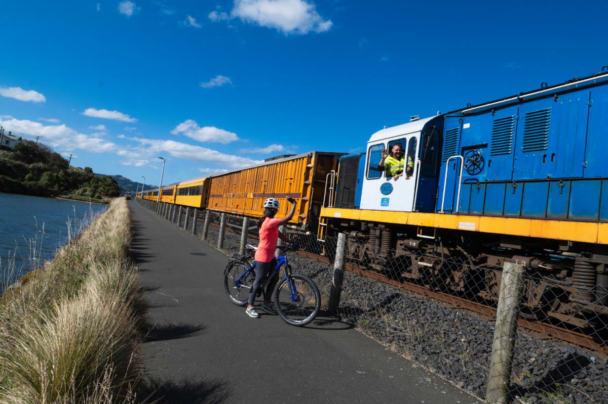 A cyclist waves at the driver of The Seasider train passing alongside a paved path.