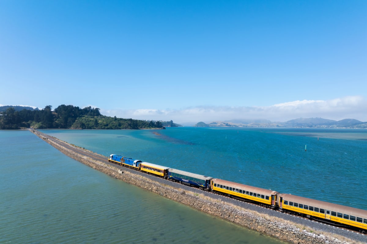 The Seasider passenger train travels along a narrow Dunedin causeway through a bright blue body of water.