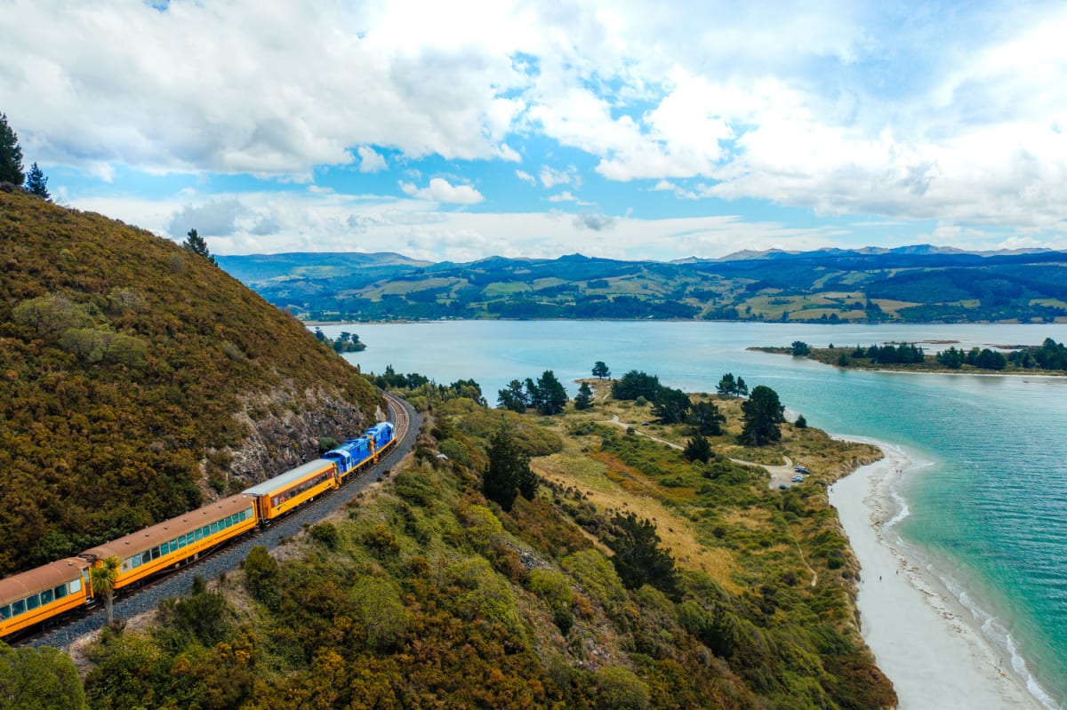 An aerial view shows The Seasider train curving along a hillside track beside the Otago blue coastal bay.