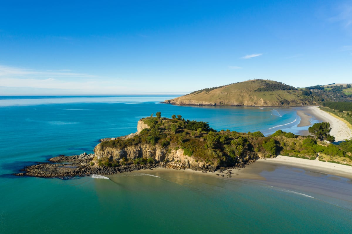 Aerial view of a rocky Dunedin, Otago NZ headland and sandy beach along a turquoise ocean under a clear blue sky.