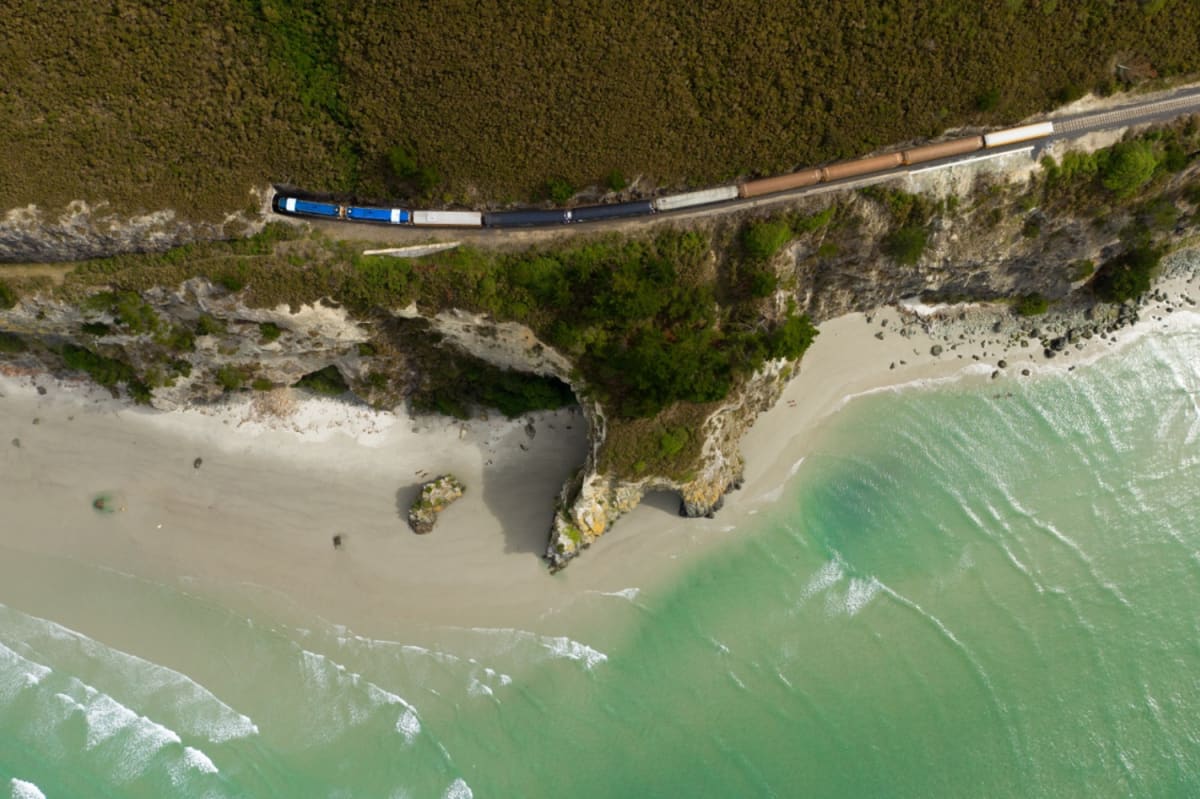 An aerial view shows the Seasider train traveling along a coastal cliff above a sandy beach and turquoise ocean waves.