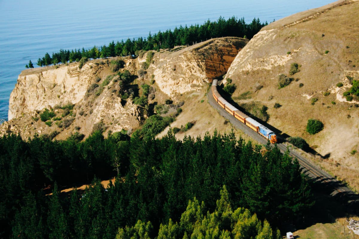 An aerial view shows the Seasider train traveling along tracks on a grassy hillside above the ocean.