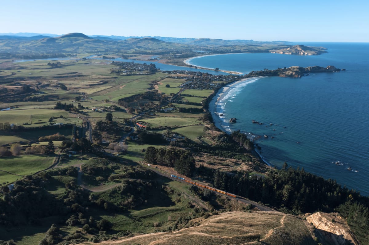 Aerial view of The Seasider train traveling along the Otago coastal railway beside green hills and blue ocean.