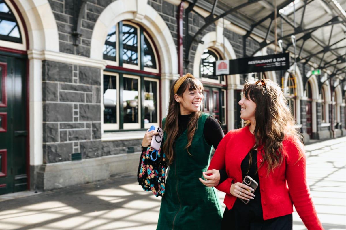 Two smiling young women walk together on a sunny outdoor train platform at the historic Dunedin Railway Station.