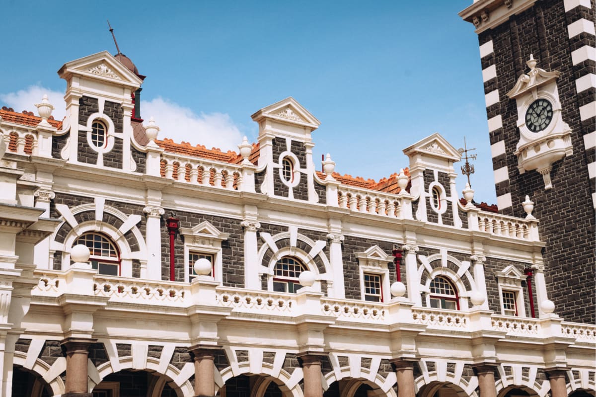 Detailed historical Dunedin Railway Station building with dark stone walls, ornate white trim, arched windows, and a corner clock tower against a blue sky.