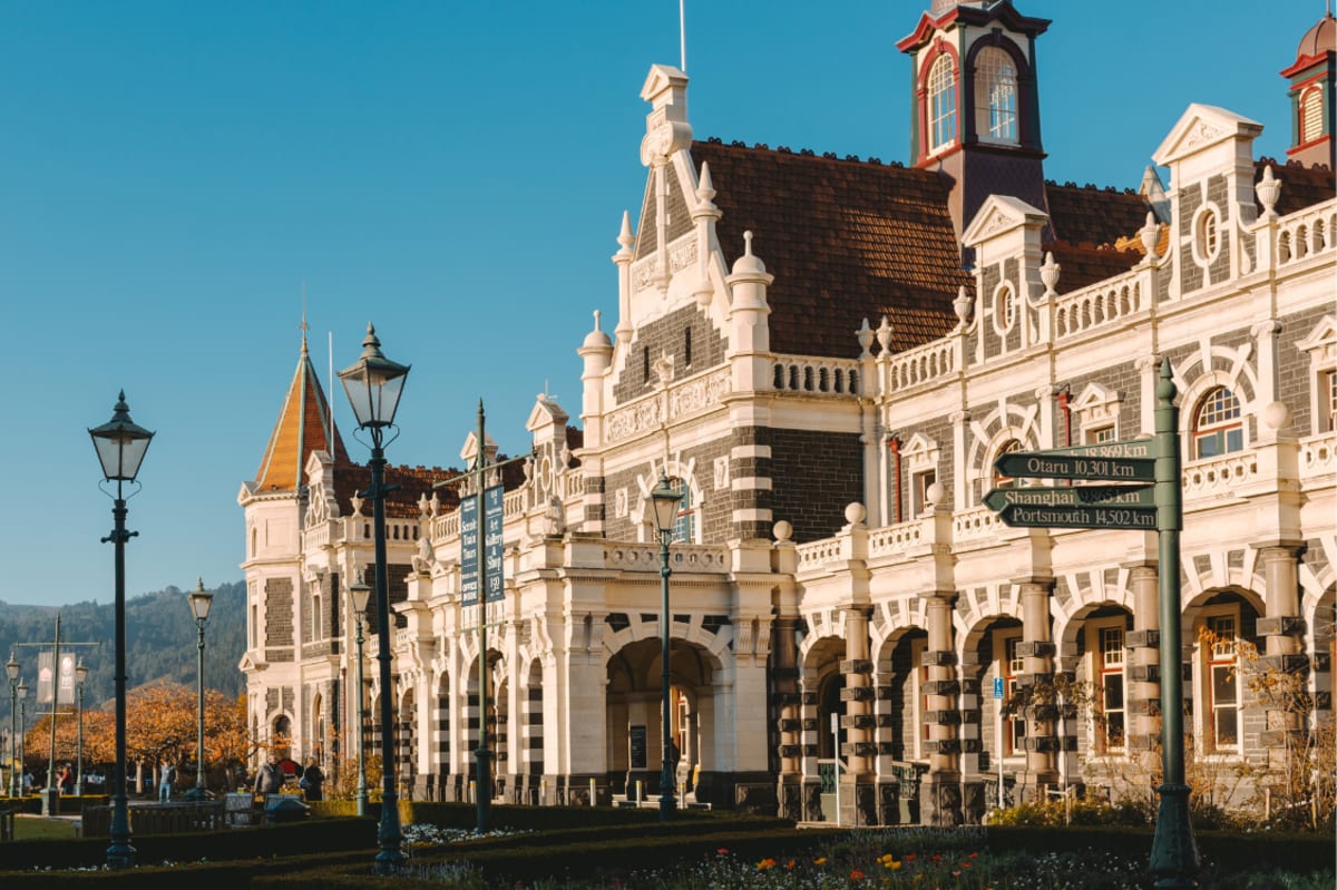The ornate stone exterior and dark brown roof of the Dunedin Railway Station against a clear blue sky.