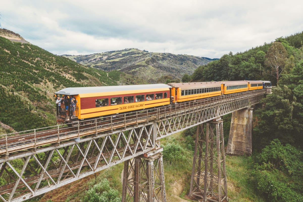 A blue and yellow train crosses a tall steel viaduct spanning a rocky, grassy gorge between rolling hills.