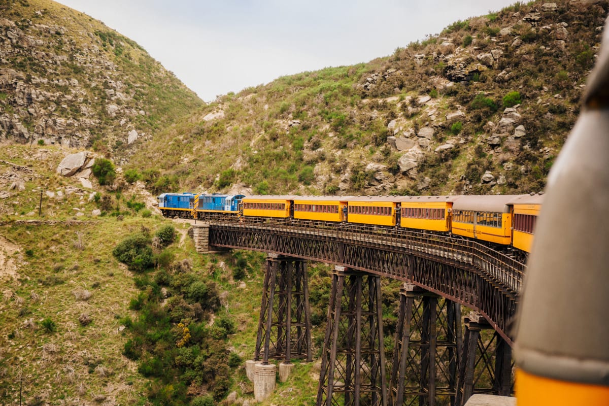 The Taieri Gorge train crosses a trestle bridge along a steep, rocky, green hillside.