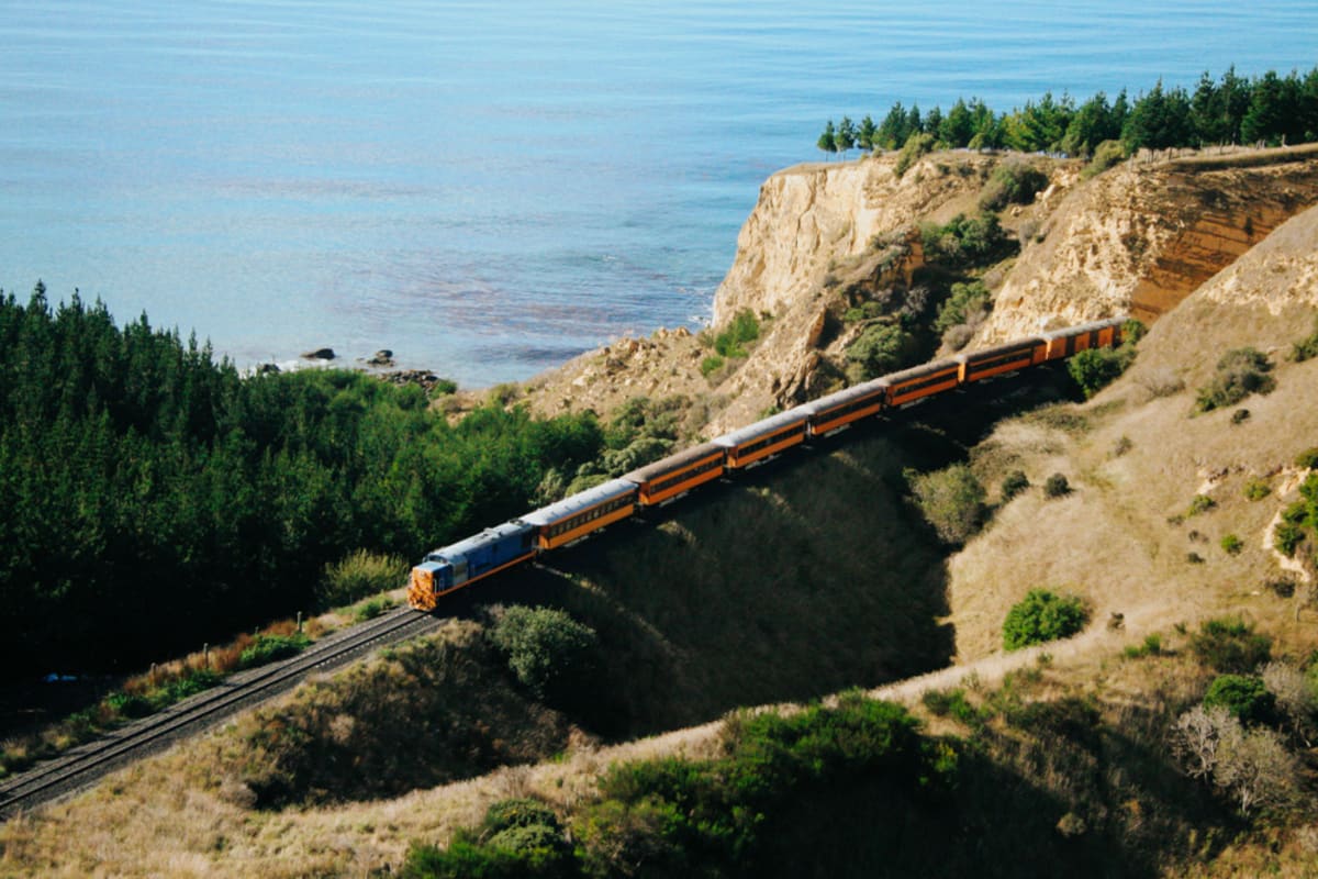 The Seasider train travels along a narrow causeway beside a body of water within Dunedin with a rolling green hill in the background.