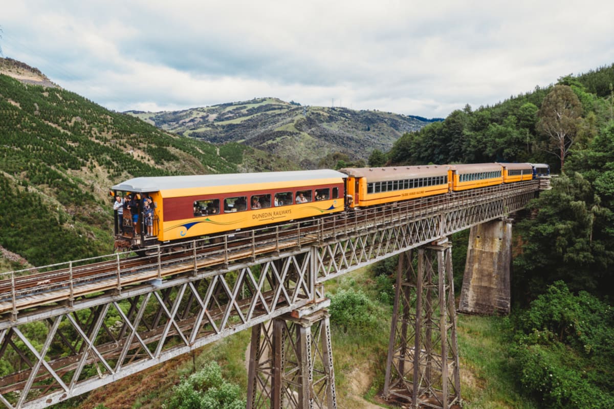 A blue and yellow train crosses a tall steel viaduct spanning a rocky, grassy gorge between rolling hills.