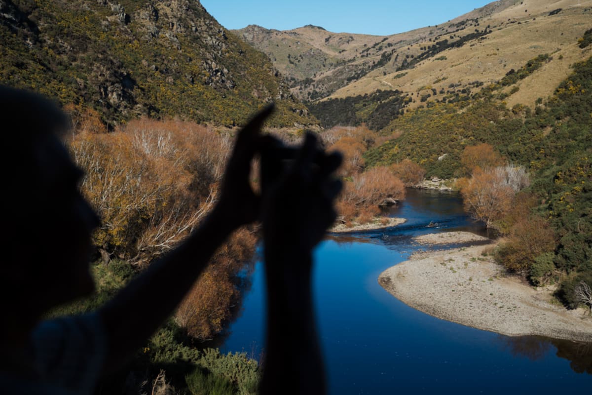 Two smiling women lean out of the Taieri Gorge train window, looking at a smartphone while traveling through a mountainous  Otago landscape.