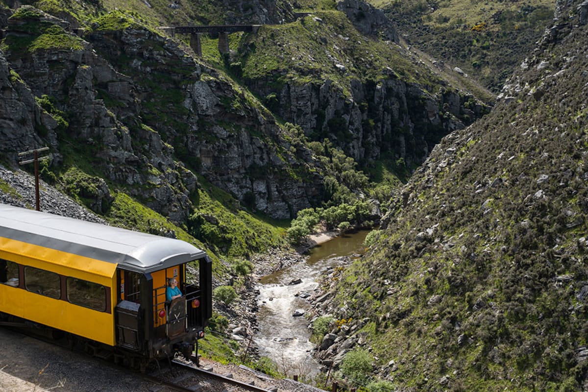 A person's feet stand on the taieri Gorge train, looking down at a steep rocky gorge and Taieri river below.