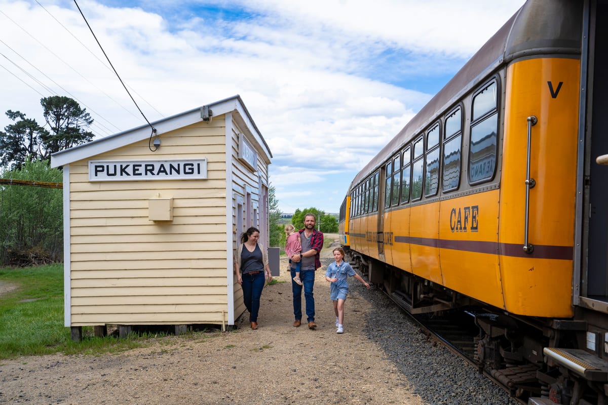 A man leans out of the Taieri Gorge train window, talking on a walkie, as people stand on the platform nearby.
