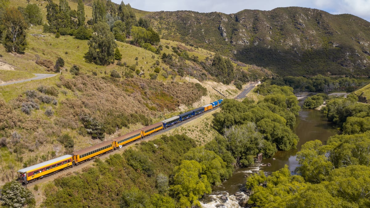 A blue and yellow train crosses a tall steel viaduct spanning a rocky, grassy gorge between rolling hills.