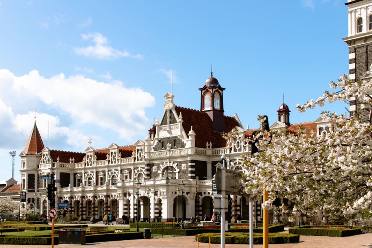 An aerial view of the historic Dunedin Railway Station in New Zealand, featuring formal gardens and a seagull flying in the foreground.