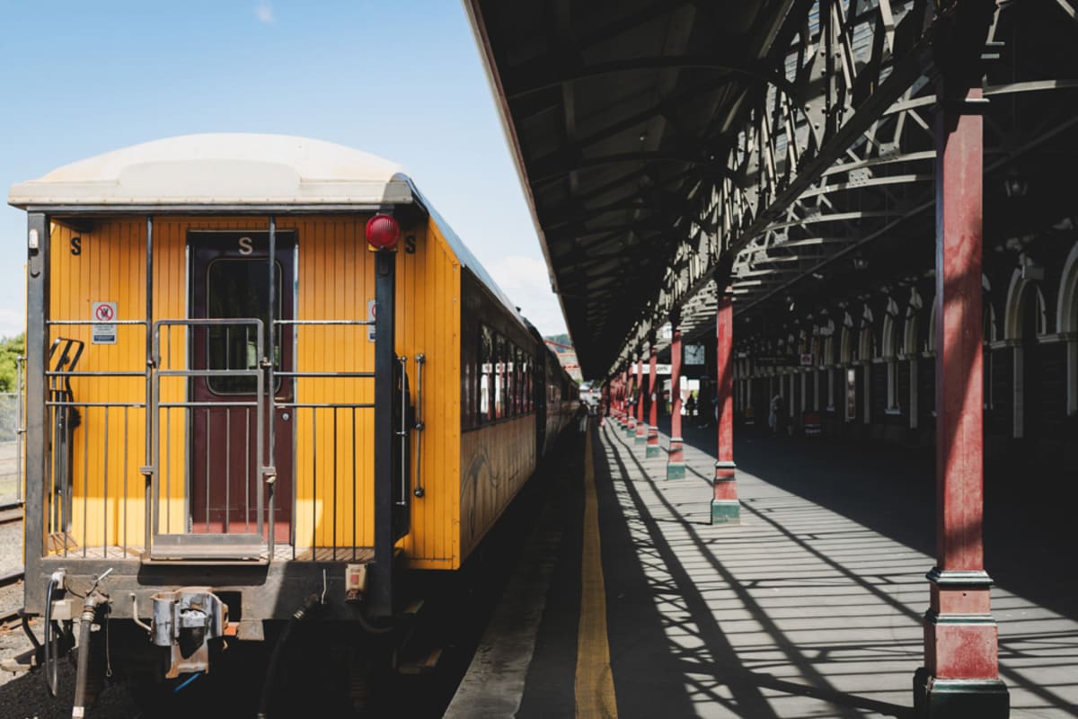 A blue and yellow train crosses a tall steel viaduct spanning a rocky, grassy gorge between rolling hills.