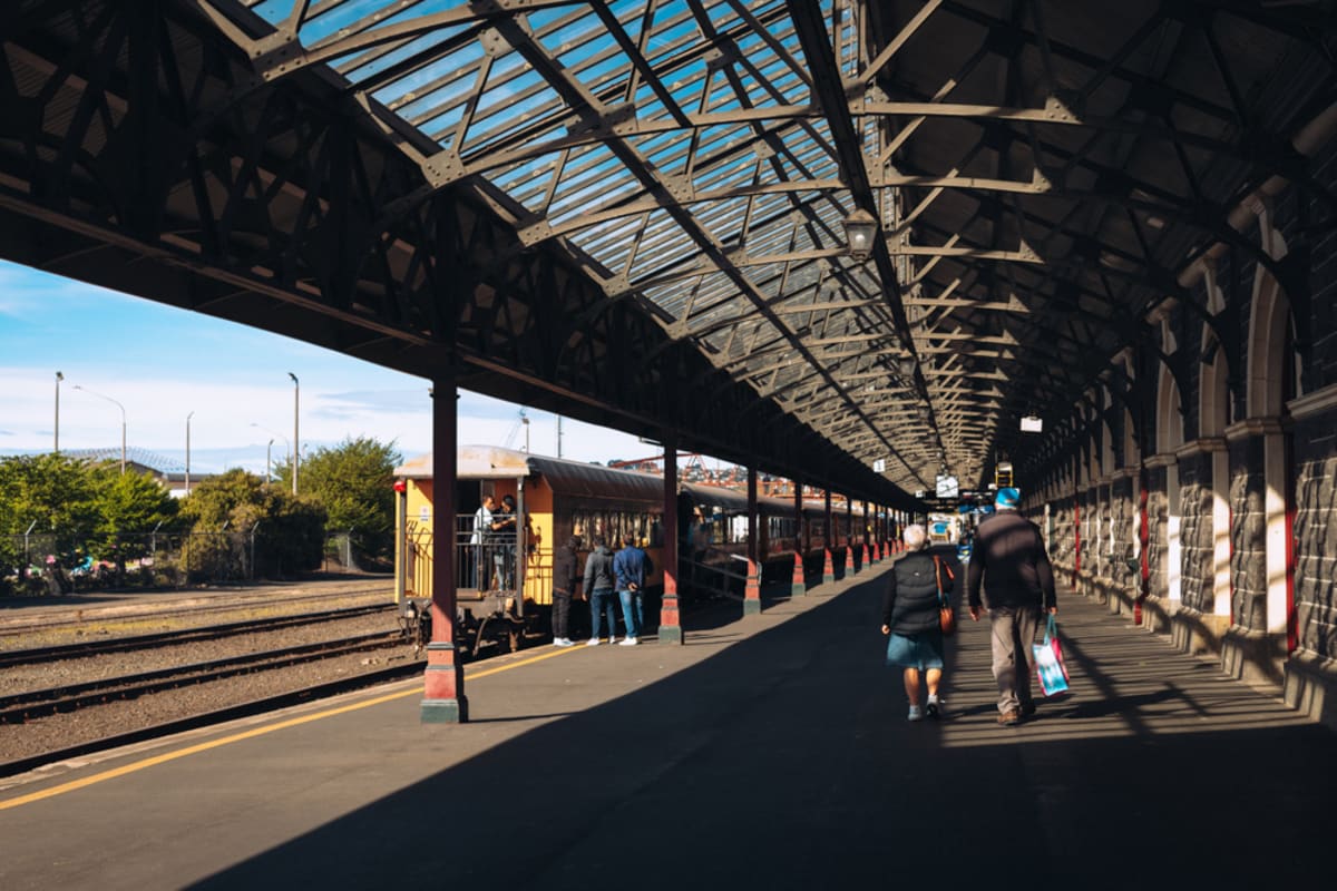 Children run along a sunlit  platform beneath a large metal roof structure and a Dunedin Railway station sign.
