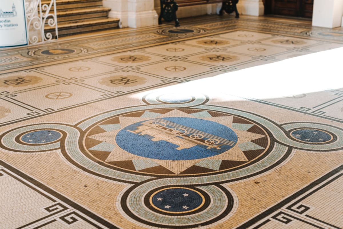 High-angle shot of an ornate yellow and white Dunedin Railway Station interior with tiled walls, arched panels, wooden benches, and a mosaic floor.