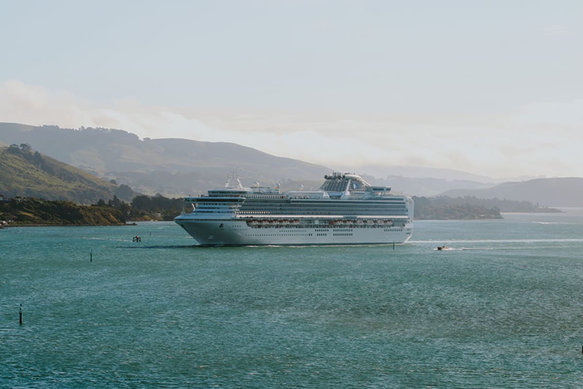 The Seasider train travels along a narrow causeway beside a body of water within Dunedin with a rolling green hill in the background.