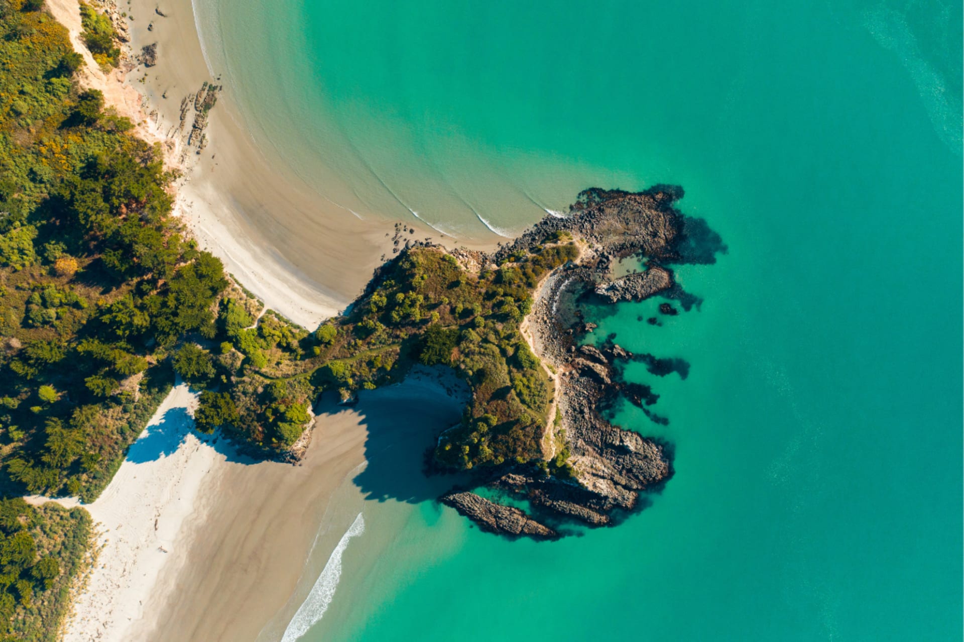 An aerial view of a sandy beach meeting bright turquoise water, with a rocky headland covered in trees extending into the sea.