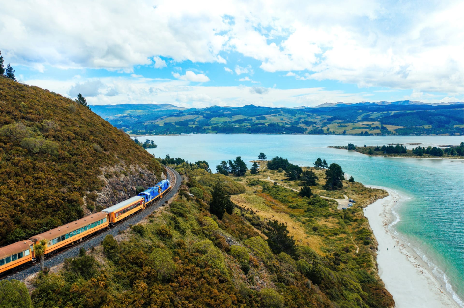 An aerial view of The Seasider train traveling along the Otago coastal track next to a blue bay and green hills.