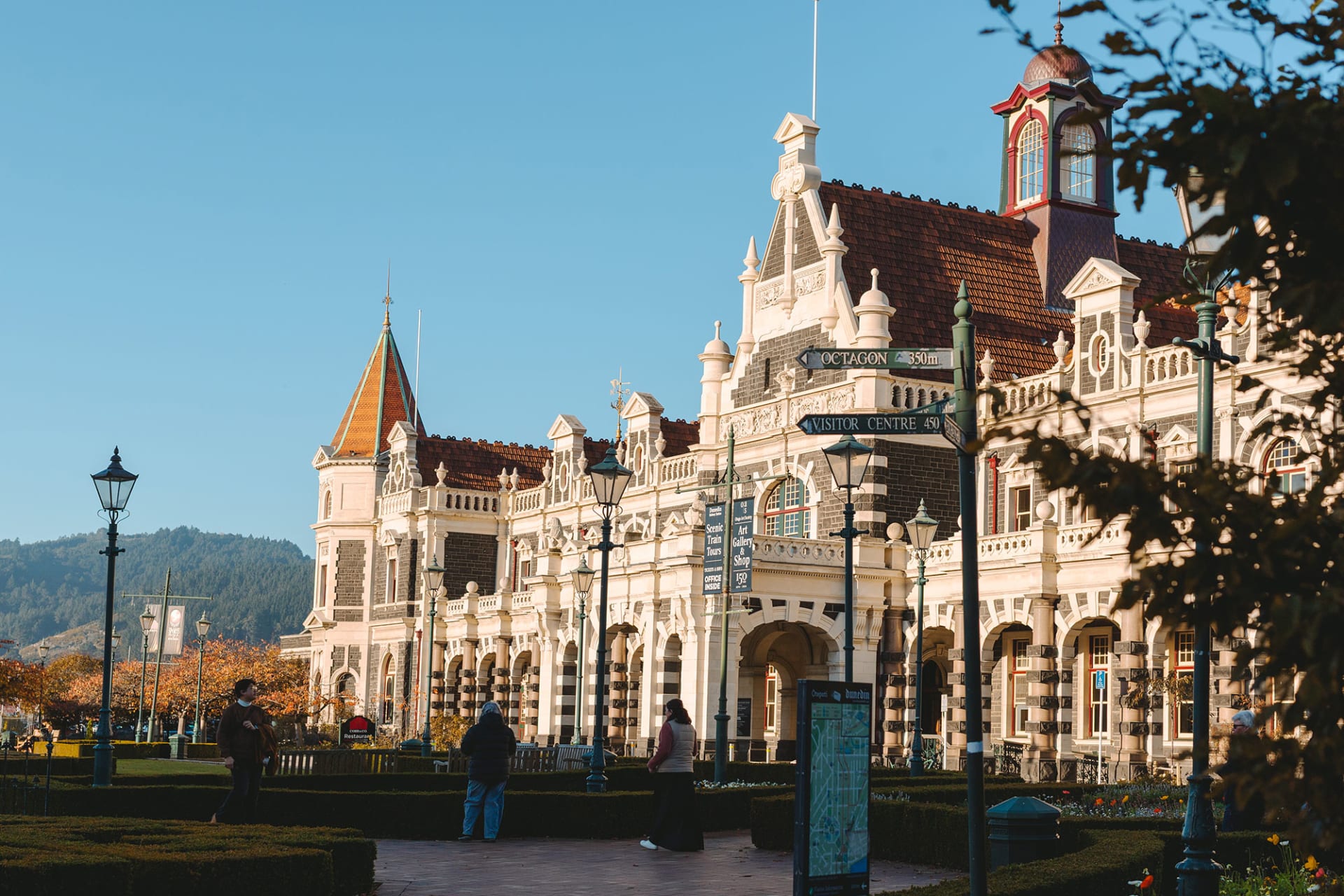 An aerial view of the grand, ornate Dunedin Railway Station surrounded by gardens, city streets, and rolling hills under a blue sky.