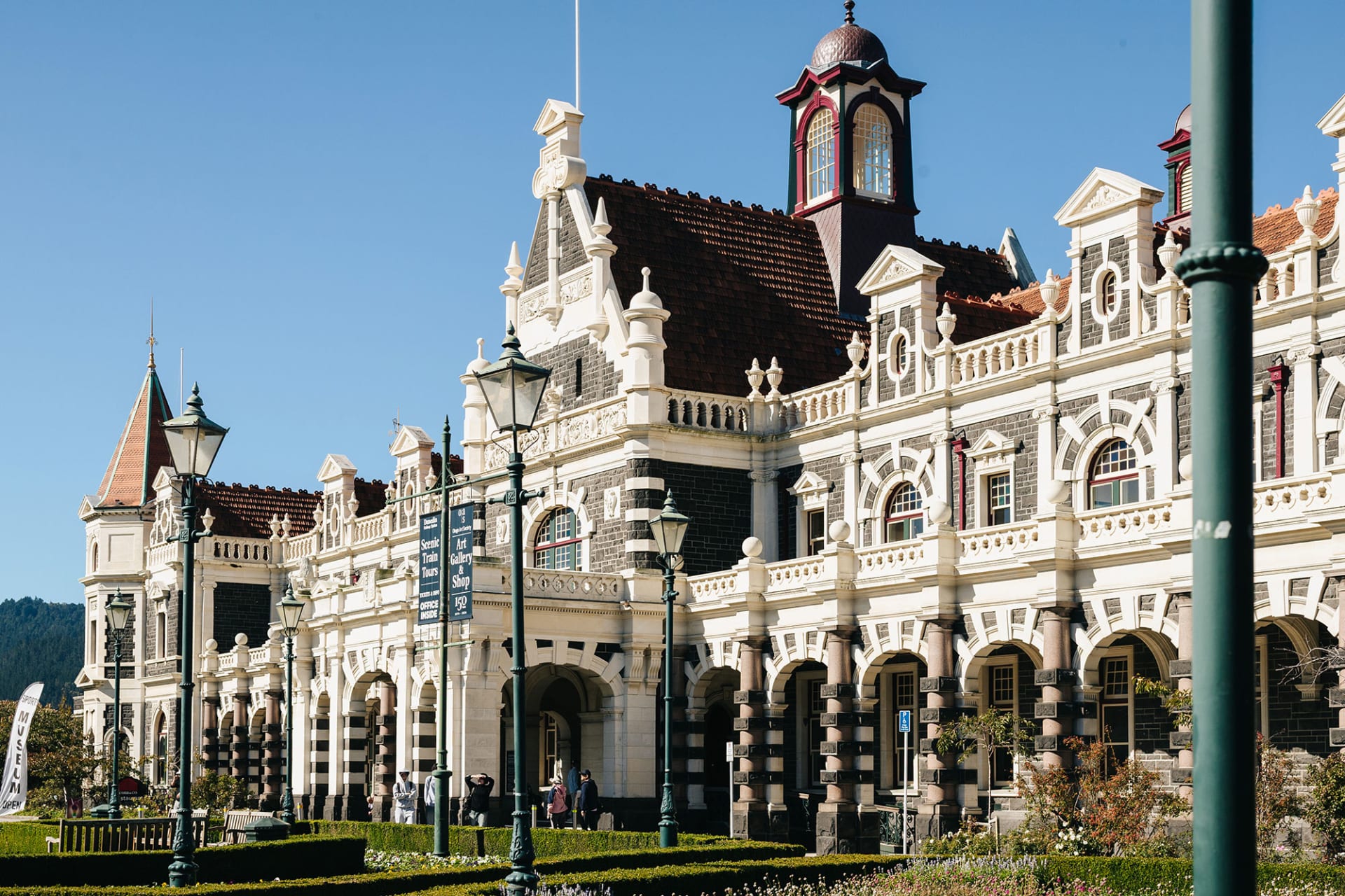 Ornate historic Dunedin Railway Station with its clock tower behind manicured gardens and blooming white cherry blossoms.