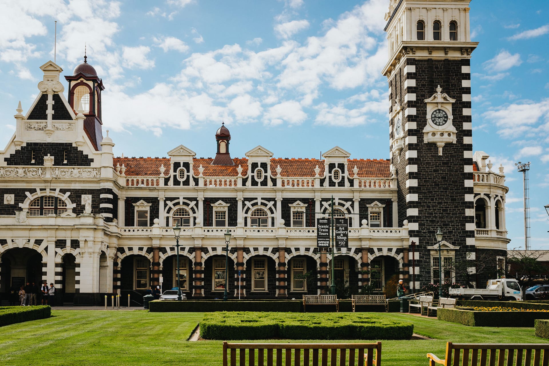 An aerial view of the historic Dunedin Railway Station in New Zealand, featuring formal gardens and a seagull flying in the foreground.