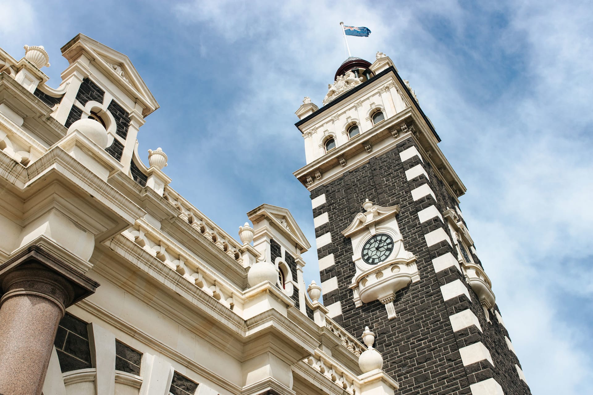An aerial view of the historic Dunedin Railway Station in New Zealand, featuring formal gardens and a seagull flying in the foreground.