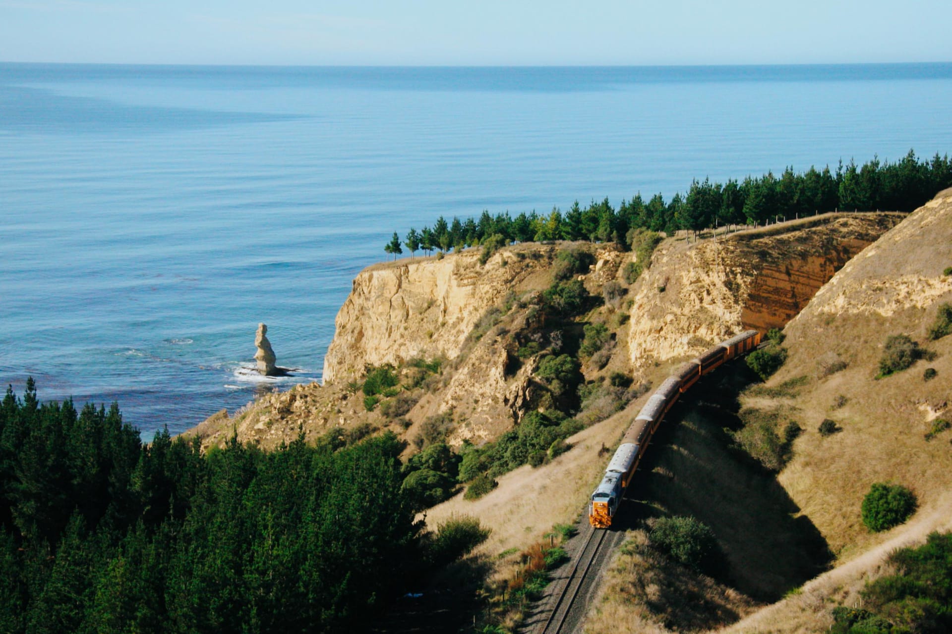 The Seasider train travels along a narrow causeway beside a body of water within Dunedin with a rolling green hill in the background.