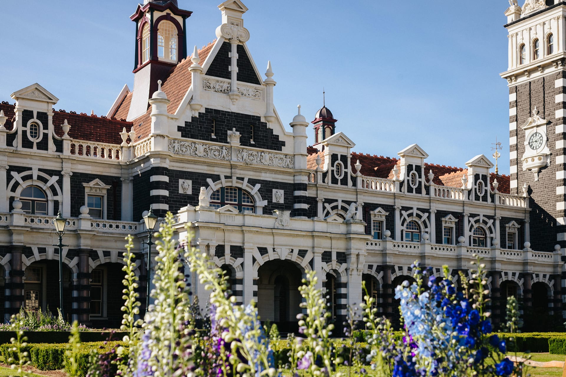 An aerial view of the historic Dunedin Railway Station in New Zealand, featuring formal gardens and a seagull flying in the foreground.