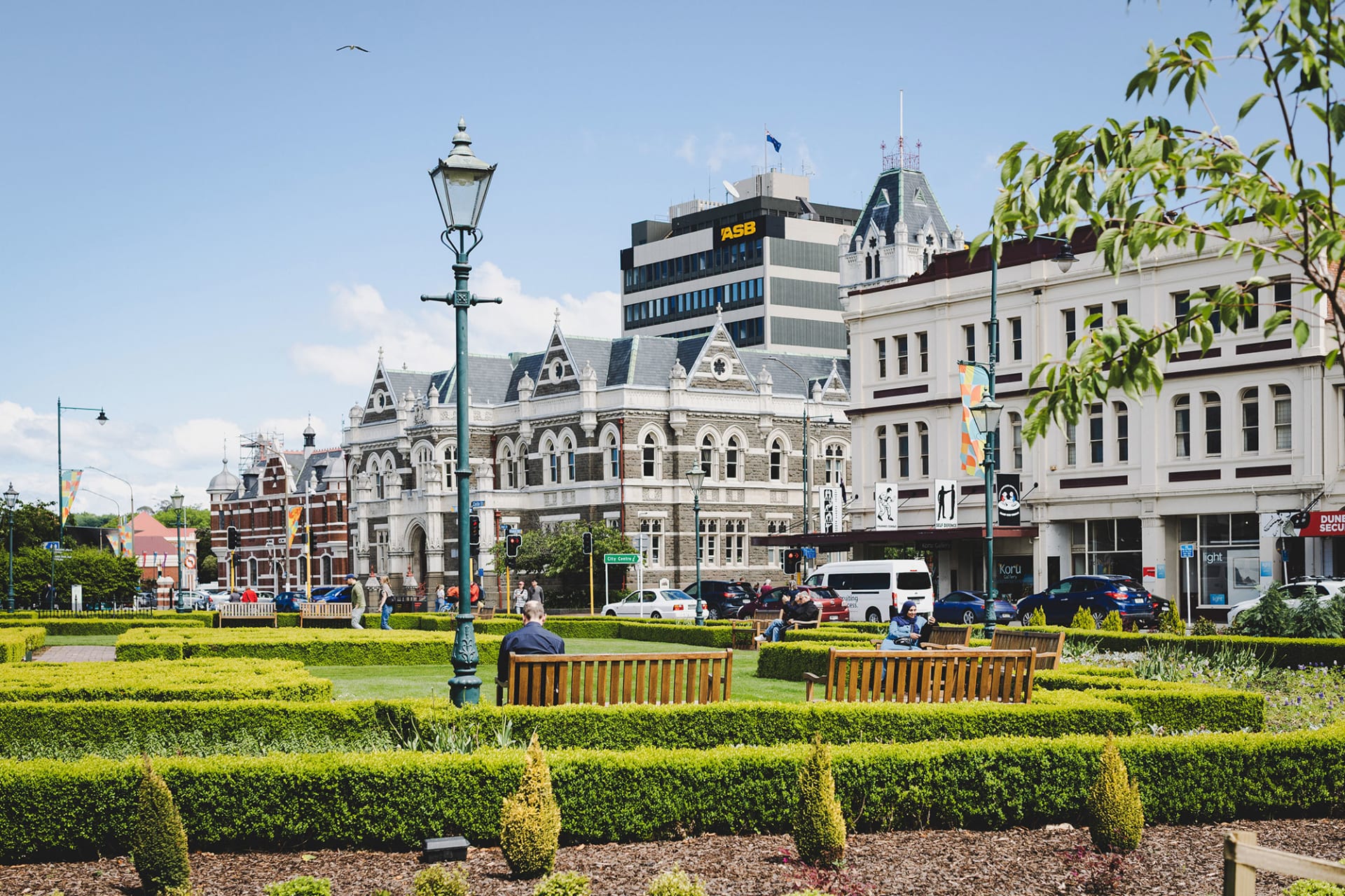 An aerial view of the historic Dunedin Railway Station in New Zealand, featuring formal gardens and a seagull flying in the foreground.