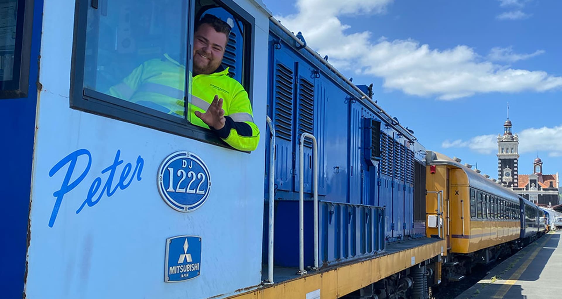 An aerial view of the grand, ornate Dunedin Railway Station surrounded by gardens, city streets, and rolling hills under a blue sky.