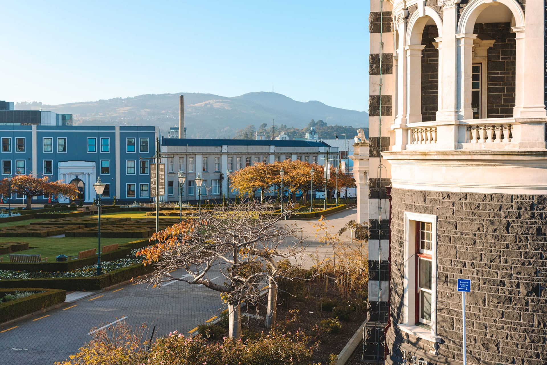 An aerial view of the Dunedin Railway Station with a manicured front garden, surrounded by Dunedin city buildings and hills under a blue sky.