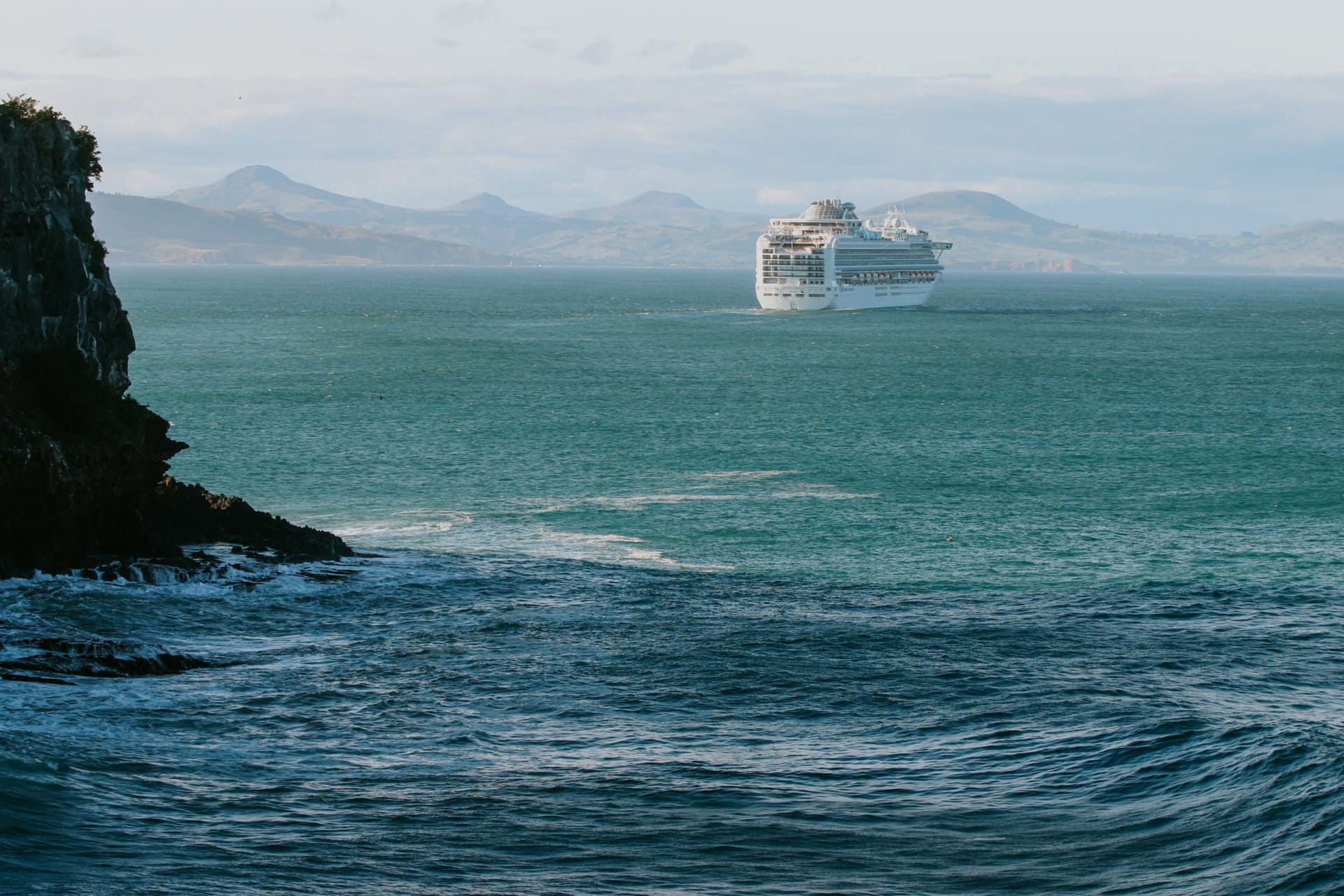 The Seasider train travels along a narrow causeway beside a body of water within Dunedin with a rolling green hill in the background.
