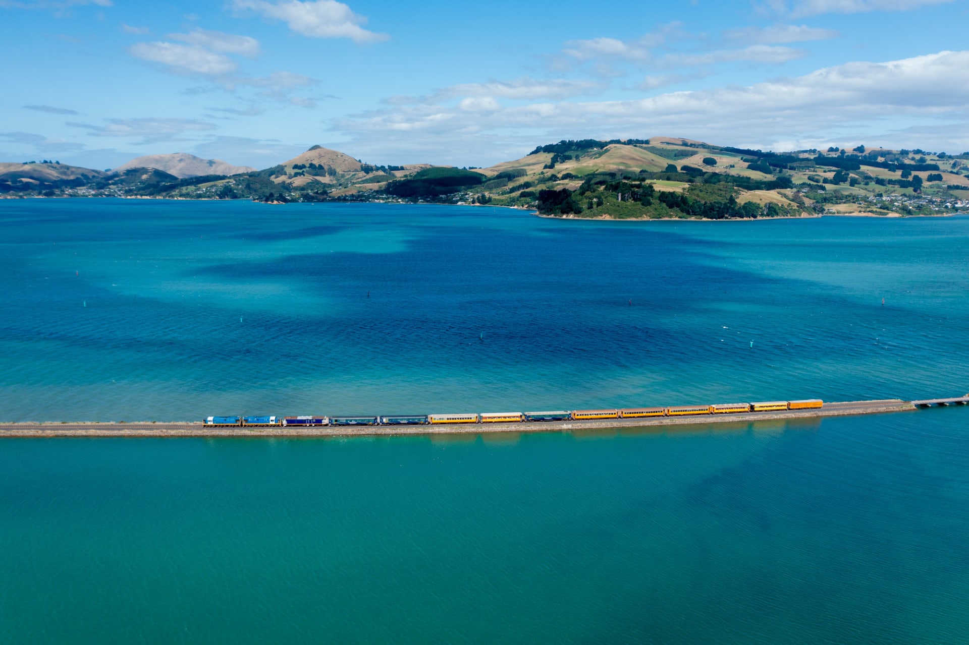 An aerial view of the grand, ornate Dunedin Railway Station surrounded by gardens, city streets, and rolling hills under a blue sky.