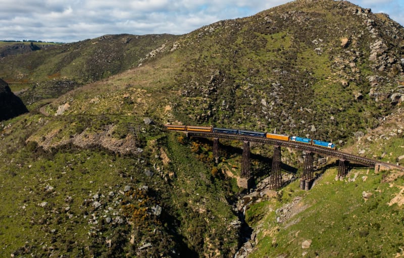 A blue and yellow train crosses a tall steel viaduct spanning a rocky, grassy gorge between rolling hills.