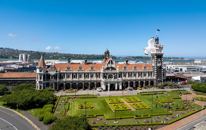 An aerial view of the historic Dunedin Railway Station in New Zealand, featuring formal gardens and a seagull flying in the foreground.