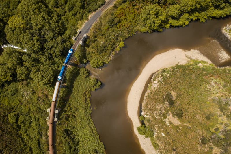 An overhead view of athe Taieri Gorge train on tracks running alongside a winding Taieri river with a sandy bank.