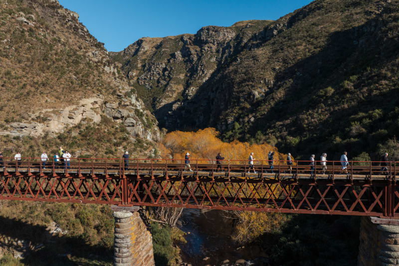 A group of people walks across a rust-colored Wingatui Viaduct bridge spanning the Taieri river in a rugged Otago mountain valley.