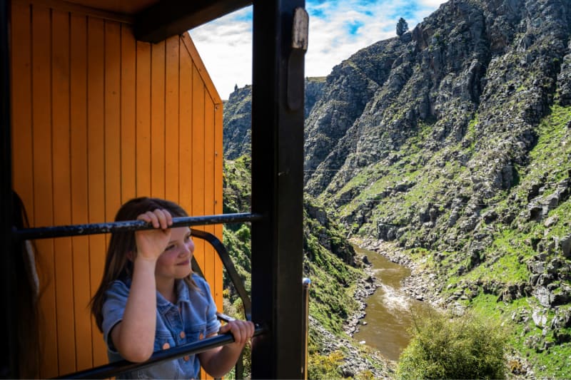 A young girl leans against a railing on the Taieri Gorge train, looking out at a rocky gorge and the Taieri river below.